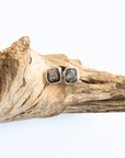 A pair of square stud earrings with iridescent opal-like centers in silver bezels, displayed on a piece of natural driftwood against a white background.