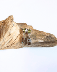 A pair of dangle earrings with peridot olive green top stones and boulder opals in silver settings, displayed on a piece of natural driftwood against a white background.