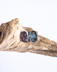 A pair of silver stud earrings with oval, iridescent opal gemstones, displayed on a piece of natural driftwood against a white background.
