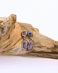 A pair of silver dangle earrings with round pink spinel blue sapphire top stones, and teardrop-shaped iridescent opal-like drops, displayed on a piece of driftwood against a white background.