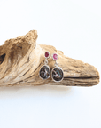 A pair of handmade dangle earrings displayed on a piece of natural driftwood against a white background. The earrings feature oval black and white marbled stones in silver bezels, with small round red gemstone studs above.