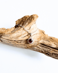 A silver chain necklace with a halo pendant featuring a round red ruby gemstone, displayed on a piece of natural driftwood against a white background.