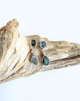 A pair of silver dangle earrings with round and teardrop-shaped iridescent green-blue opal stones, displayed on a piece of natural driftwood against a white background.