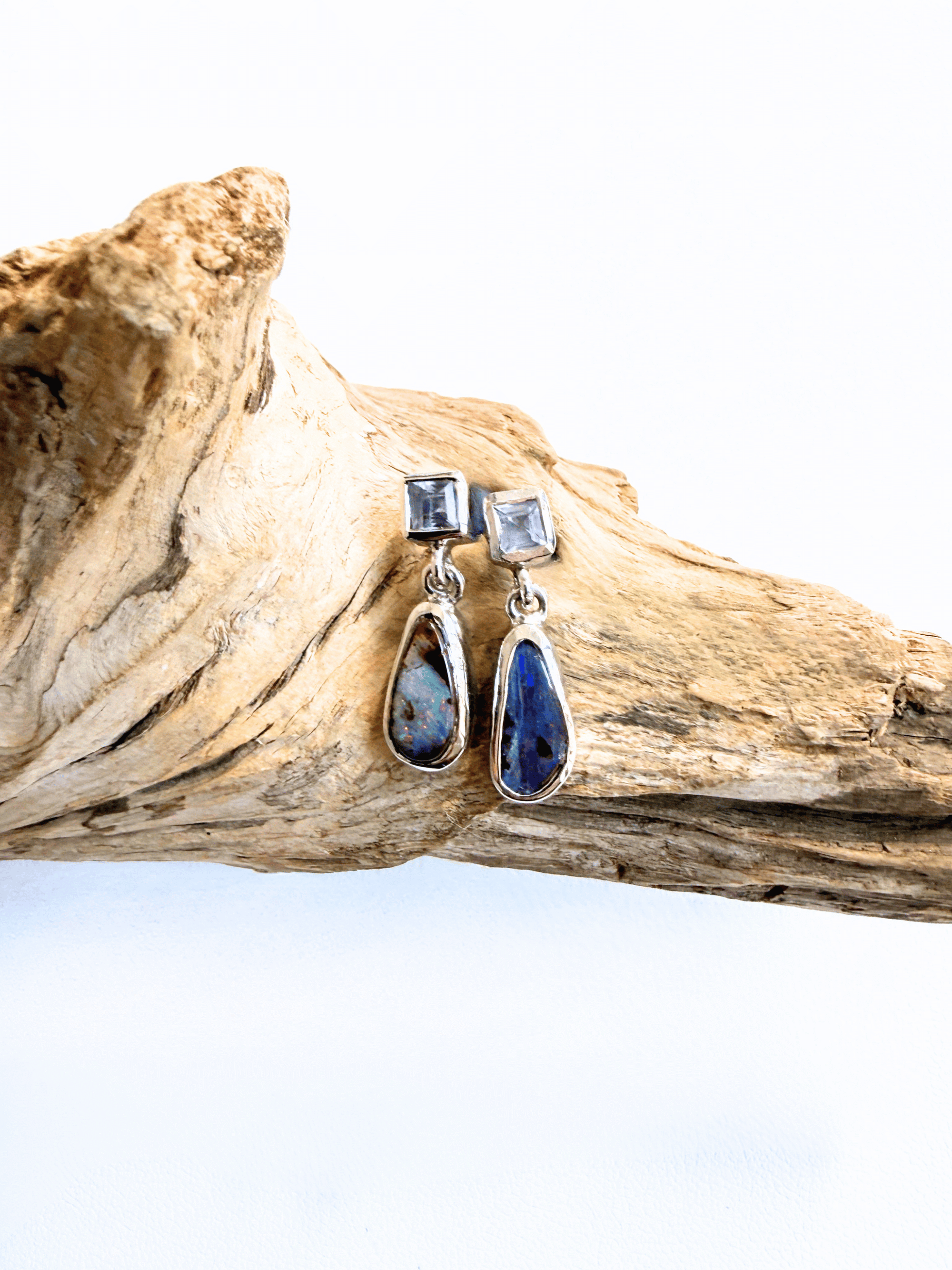 A pair of silver drop earrings with square pale blue stones at the top and teardrop-shaped dark blue and white marbled stones below, displayed on a piece of driftwood against a white background.