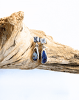 A pair of silver drop earrings with square pale blue stones at the top and teardrop-shaped dark blue and white marbled stones below, displayed on a piece of driftwood against a white background.