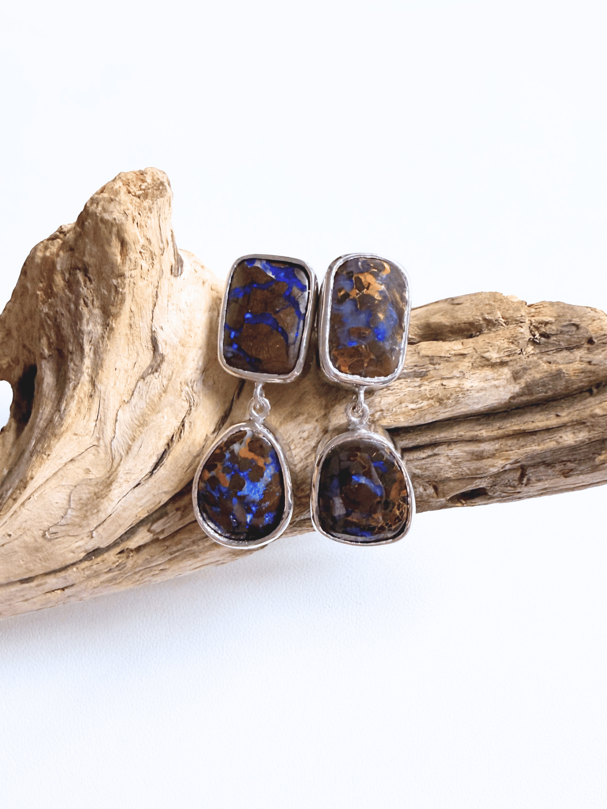 A pair of silver drop earrings with rectangular and teardrop-shaped brown and blue opal stones, displayed on a piece of natural driftwood against a white background.
