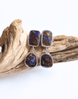 A pair of silver drop earrings with rectangular and teardrop-shaped brown and blue opal stones, displayed on a piece of natural driftwood against a white background.