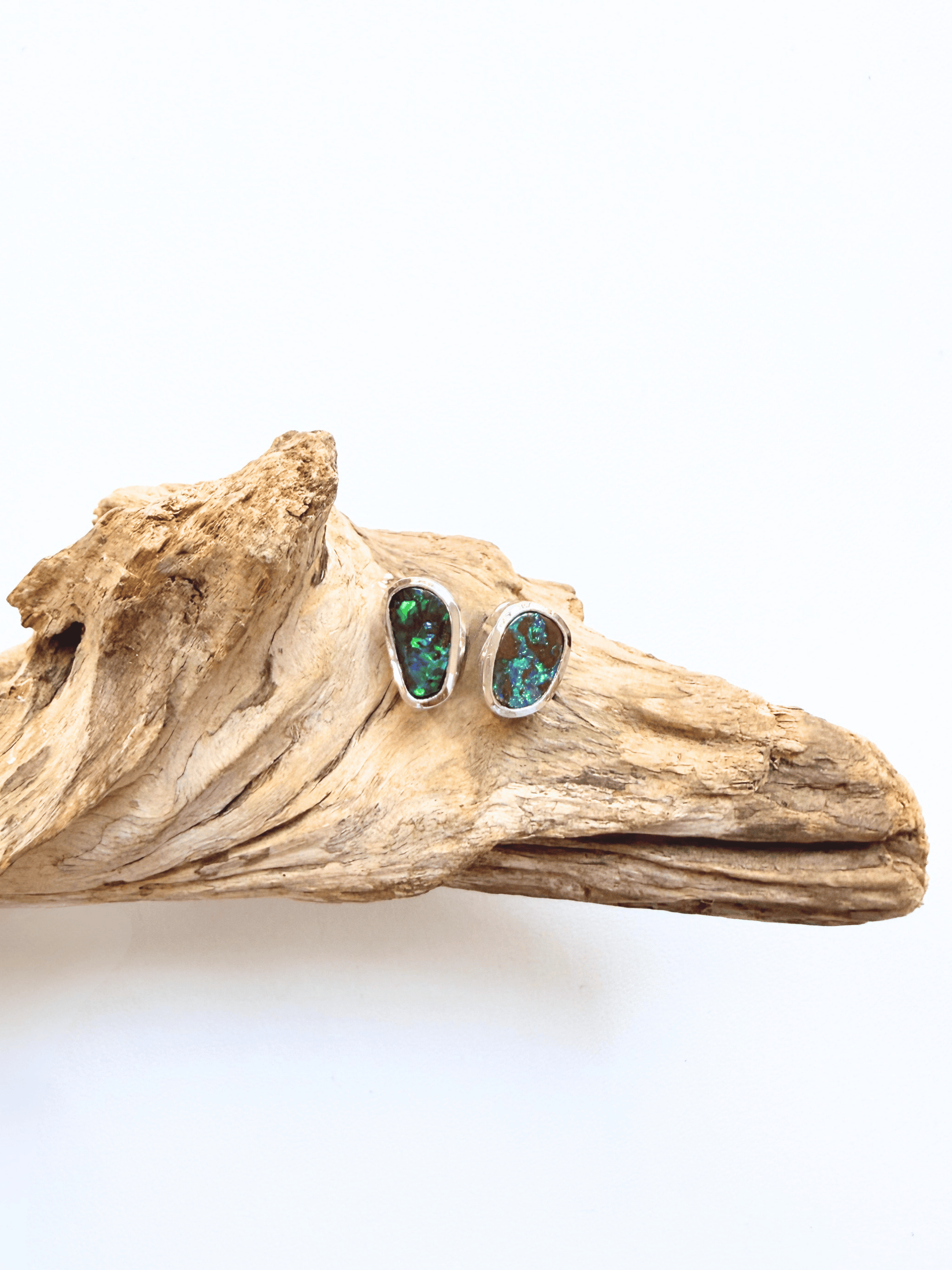 A pair of silver stud earrings with green opal inlays, displayed on a piece of natural driftwood against a plain white background.