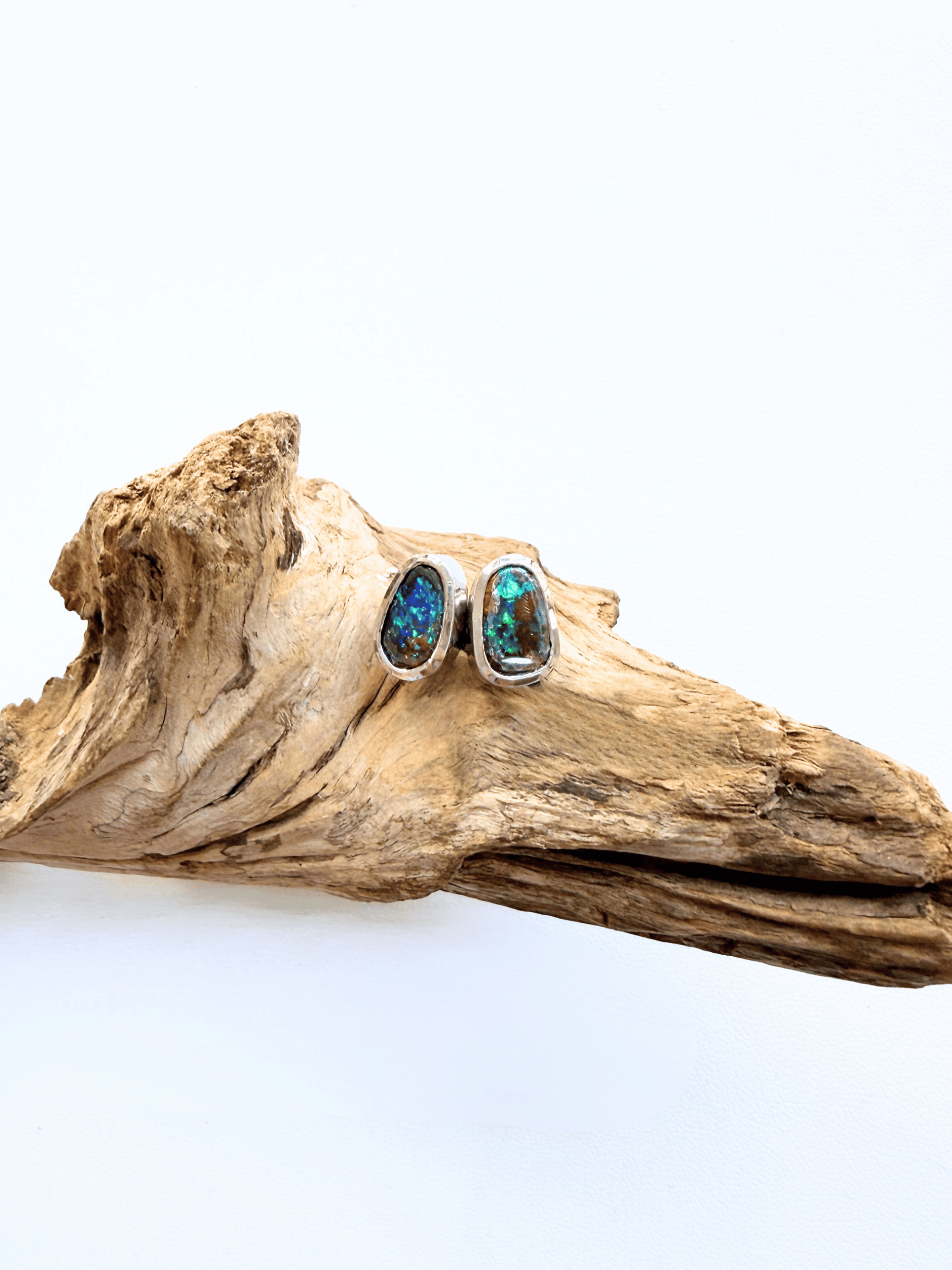 A pair of silver stud earrings with oval blue opal gemstones, displayed on a piece of natural driftwood against a plain white background.