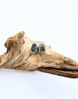 A pair of silver stud earrings with oval blue opal gemstones, displayed on a piece of natural driftwood against a plain white background.