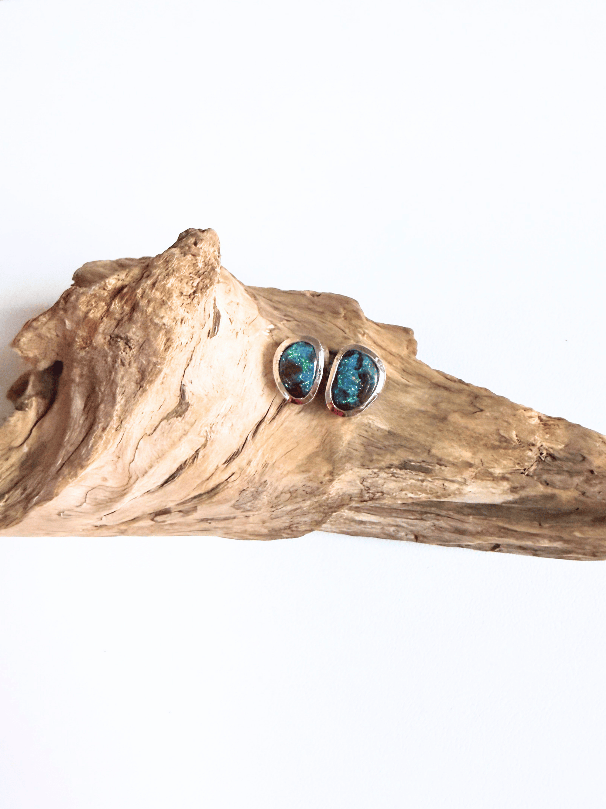 A pair of silver stud earrings with oval turquoise-blue stones, displayed on a piece of natural driftwood against a plain white background.
