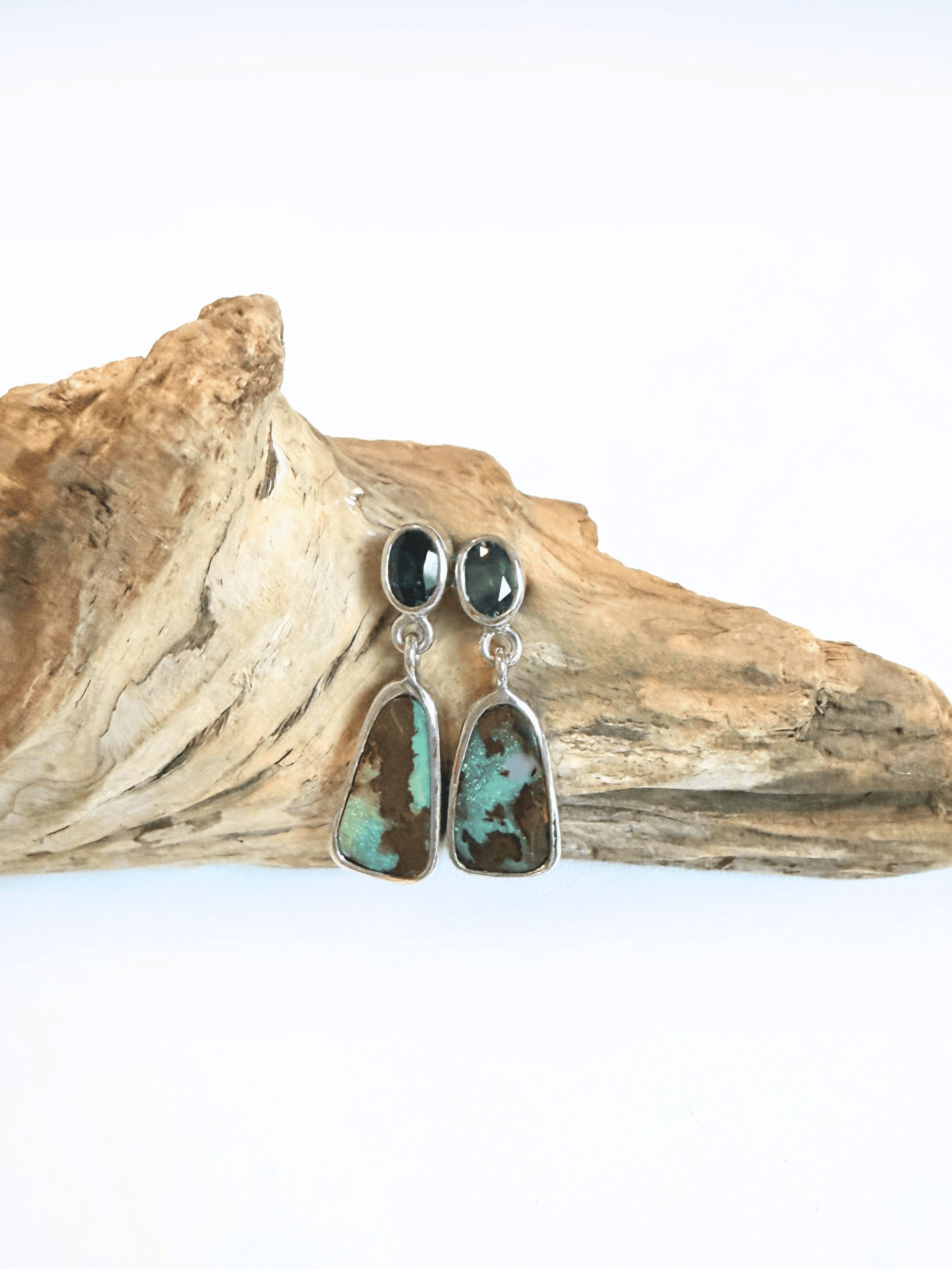 A pair of dangle earrings with dark blue round sapphires and irregular turquoise-and-brown marbled opal drops in silver settings, displayed on a piece of driftwood against a white background.