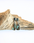 A pair of dangle earrings with dark blue round sapphires and irregular turquoise-and-brown marbled opal drops in silver settings, displayed on a piece of driftwood against a white background.