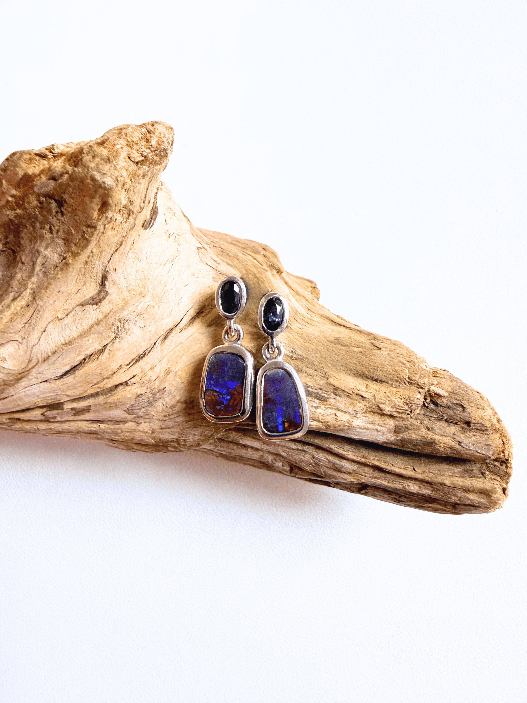 A pair of dangle earrings with dark blue sapphires to teardrop-shaped opal stones in silver settings, displayed on a piece of natural driftwood against a white background.