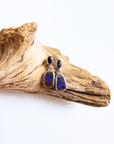 A pair of dangle earrings with dark blue sapphires to teardrop-shaped opal stones in silver settings, displayed on a piece of natural driftwood against a white background.