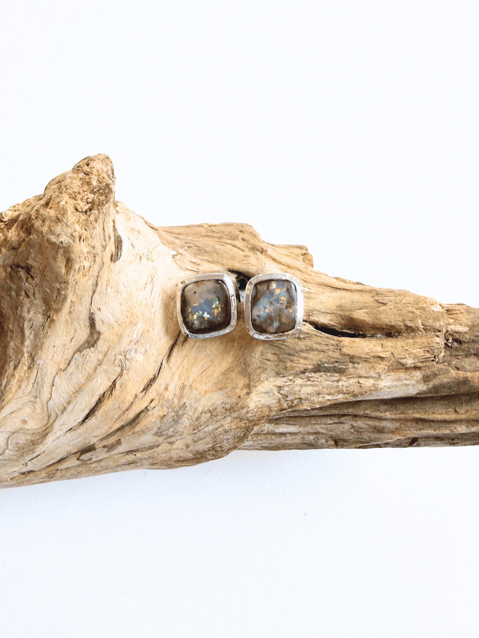 A pair of square stud earrings with iridescent opal-like centers in silver bezels, displayed on a piece of natural driftwood against a white background.