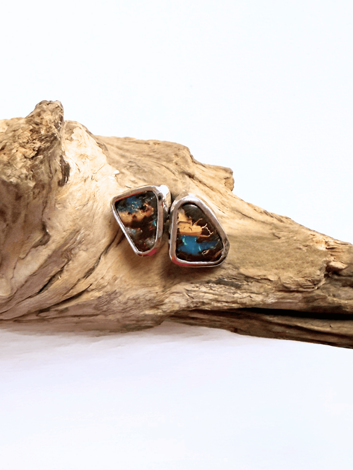 A pair of silver stud earrings with irregular triangular turquoise inlay, resting on a piece of natural driftwood against a white background.