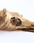 A pair of silver stud earrings with irregular triangular turquoise inlay, resting on a piece of natural driftwood against a white background.