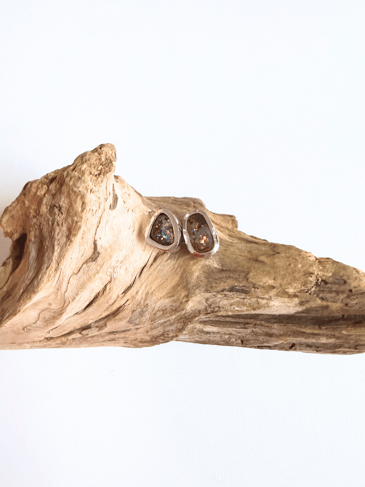 A pair of silver-toned teardrop stud earrings with iridescent glitter inlays, displayed on a piece of natural driftwood against a plain white background.