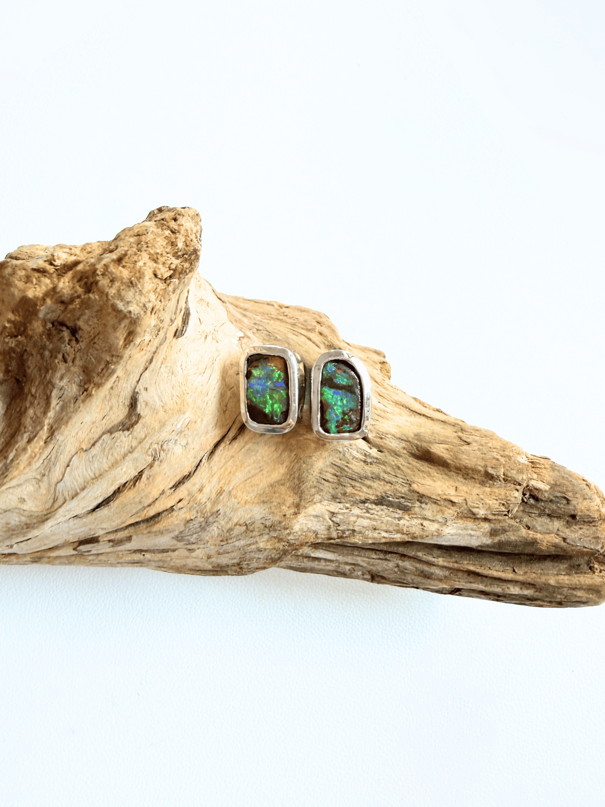 A pair of rectangular silver stud earrings with iridescent green and blue opal inlays, displayed on a piece of natural driftwood against a white background.