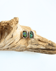 A pair of rectangular silver stud earrings with iridescent green and blue opal inlays, displayed on a piece of natural driftwood against a white background.