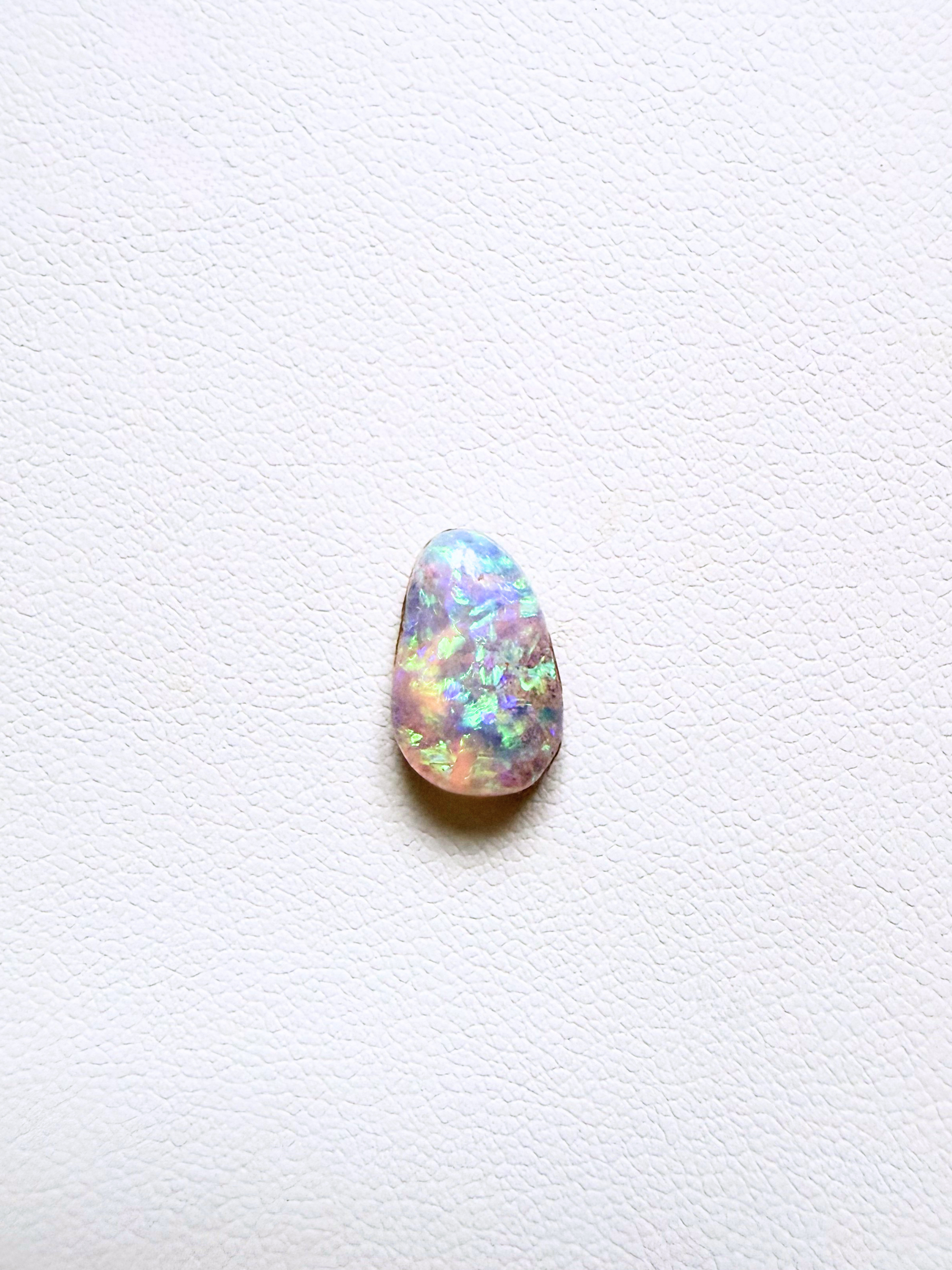A teardrop-shaped cabochon opal gemstone displaying vibrant iridescent flashes of blue, green, orange, and pink on a textured white background.
