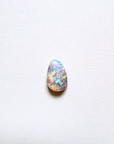 A teardrop-shaped cabochon opal gemstone displaying vibrant iridescent flashes of blue, green, orange, and pink on a textured white background.