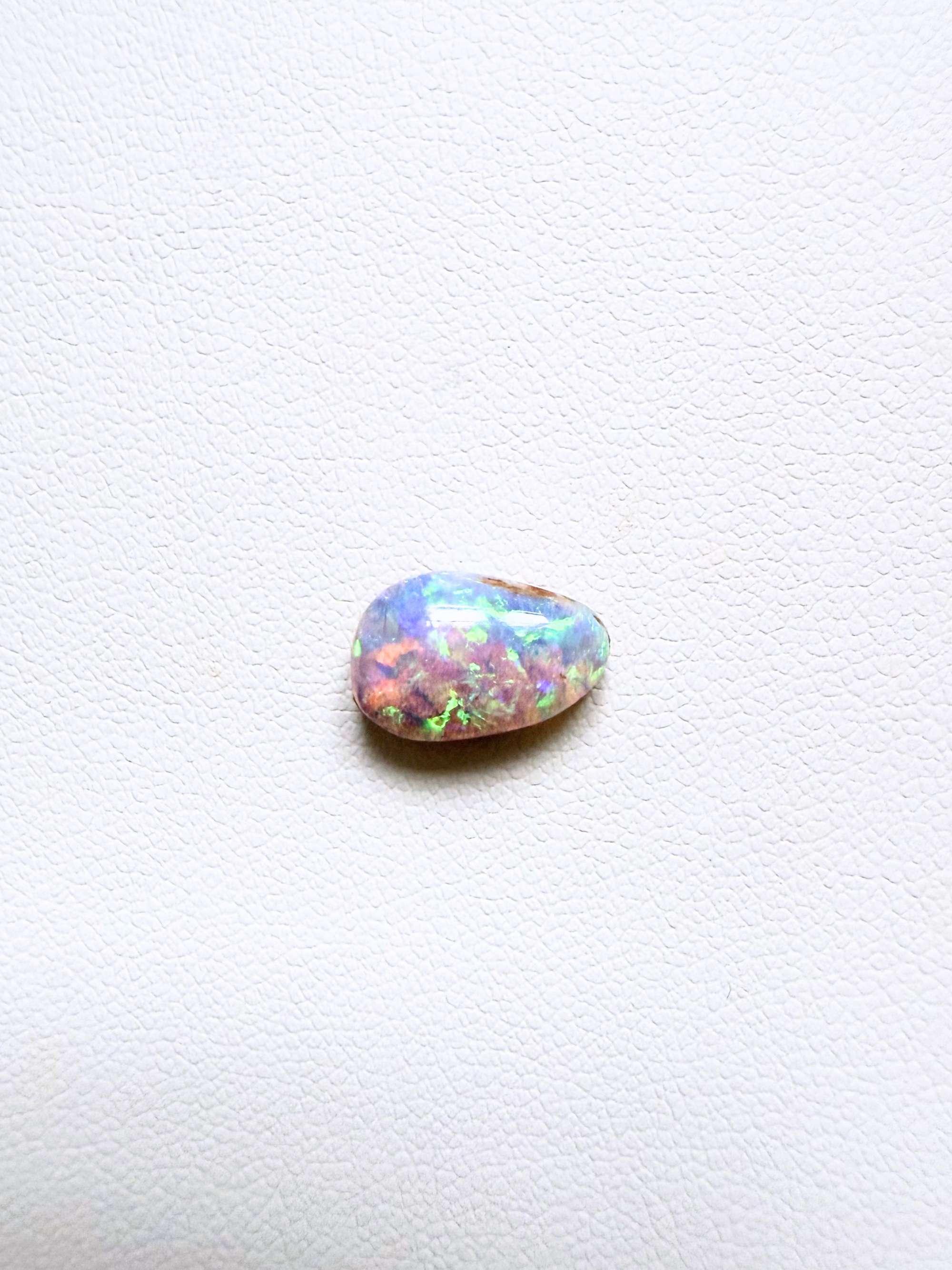A small teardrop-shaped opal gemstone with iridescent colors of blue, green, pink, and orange, resting on a textured white surface.