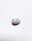 A small teardrop-shaped opal gemstone with iridescent colors of blue, green, pink, and orange, resting on a textured white surface.