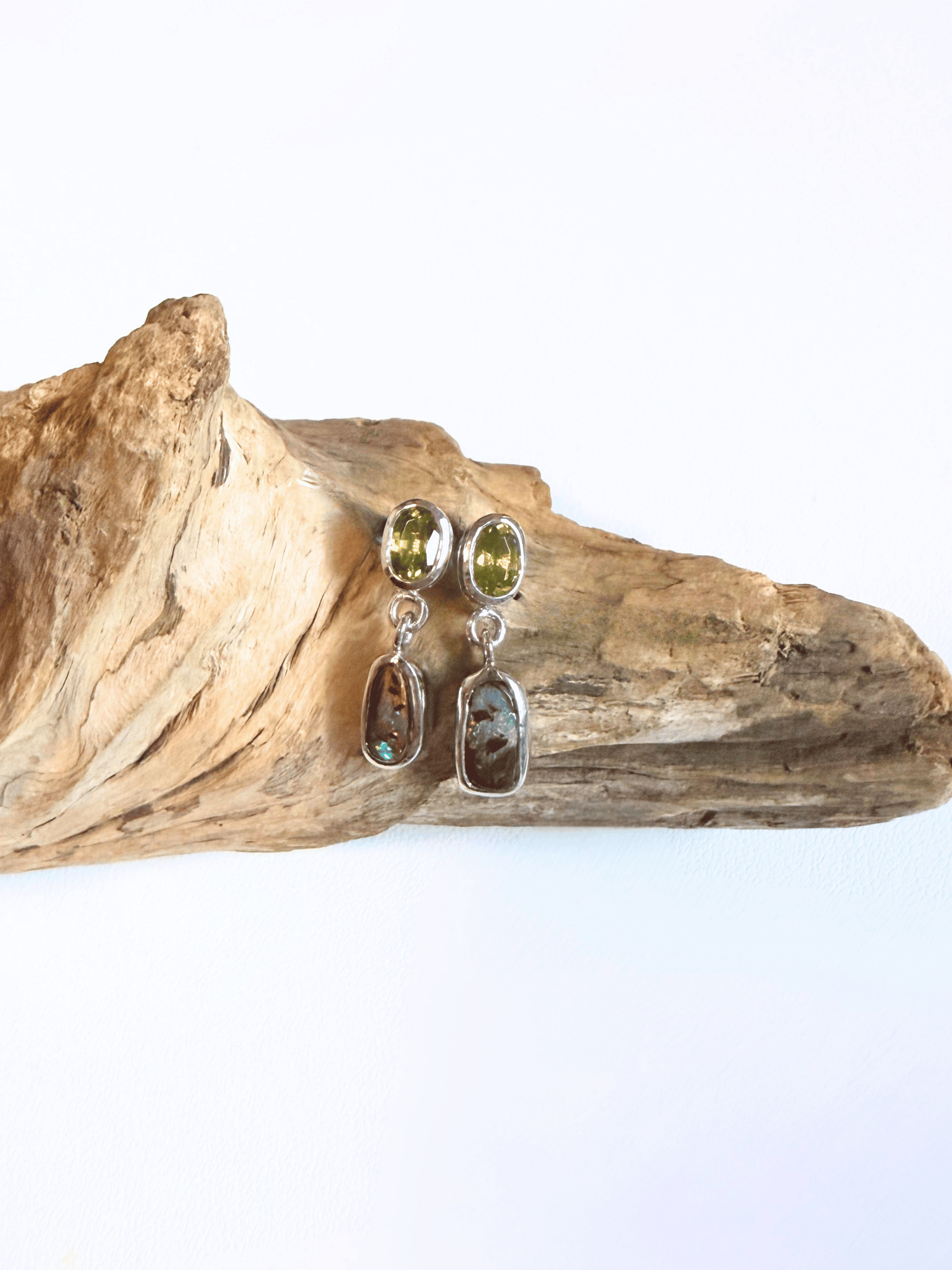 A pair of dangle earrings with peridot olive green top stones and boulder opals in silver settings, displayed on a piece of natural driftwood against a white background.