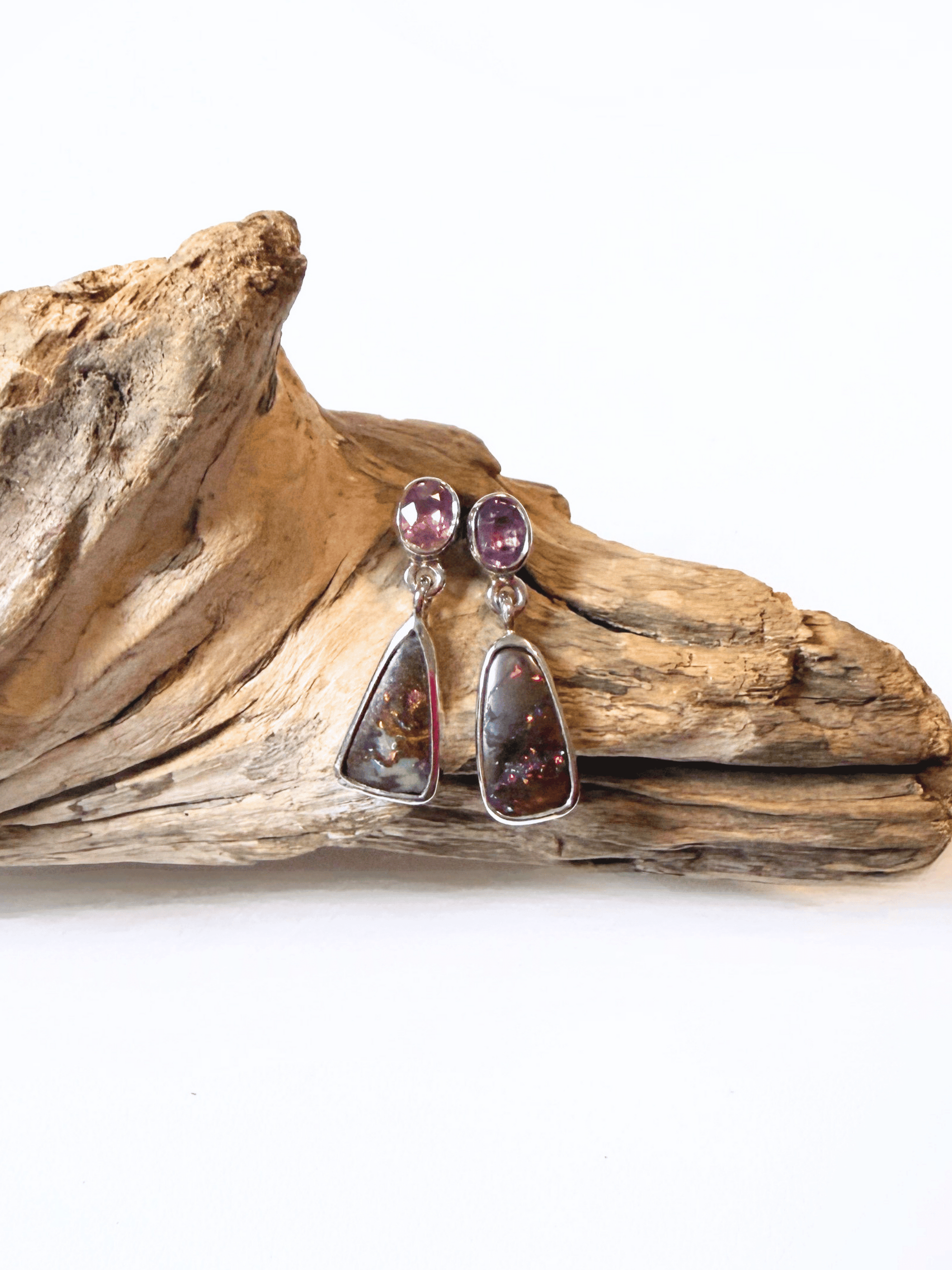 A pair of dangle earrings with oval pink sapphire top stones and teardrop-shaped boulder opal stones in silver settings, displayed on a piece of natural driftwood against a white background.