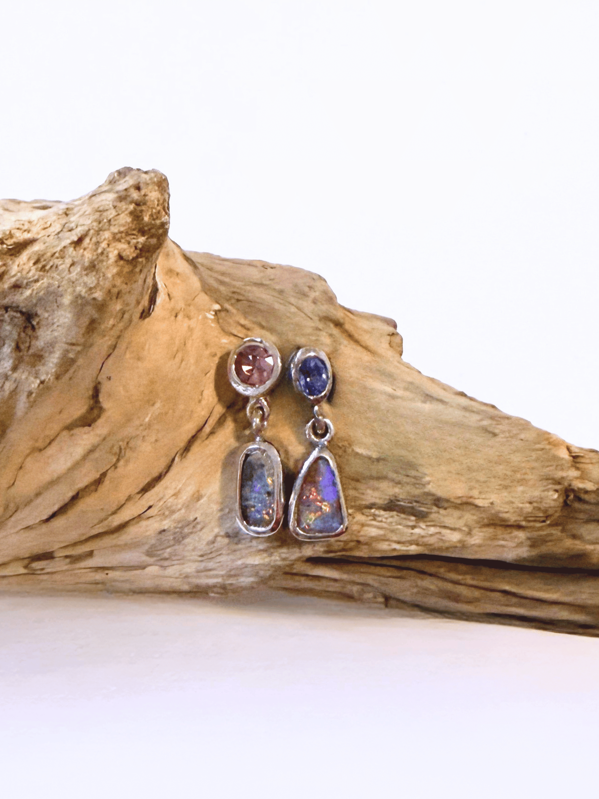 A pair of silver dangle earrings with round pink spinel blue sapphire top stones, and teardrop-shaped iridescent opal-like drops, displayed on a piece of driftwood against a white background.
