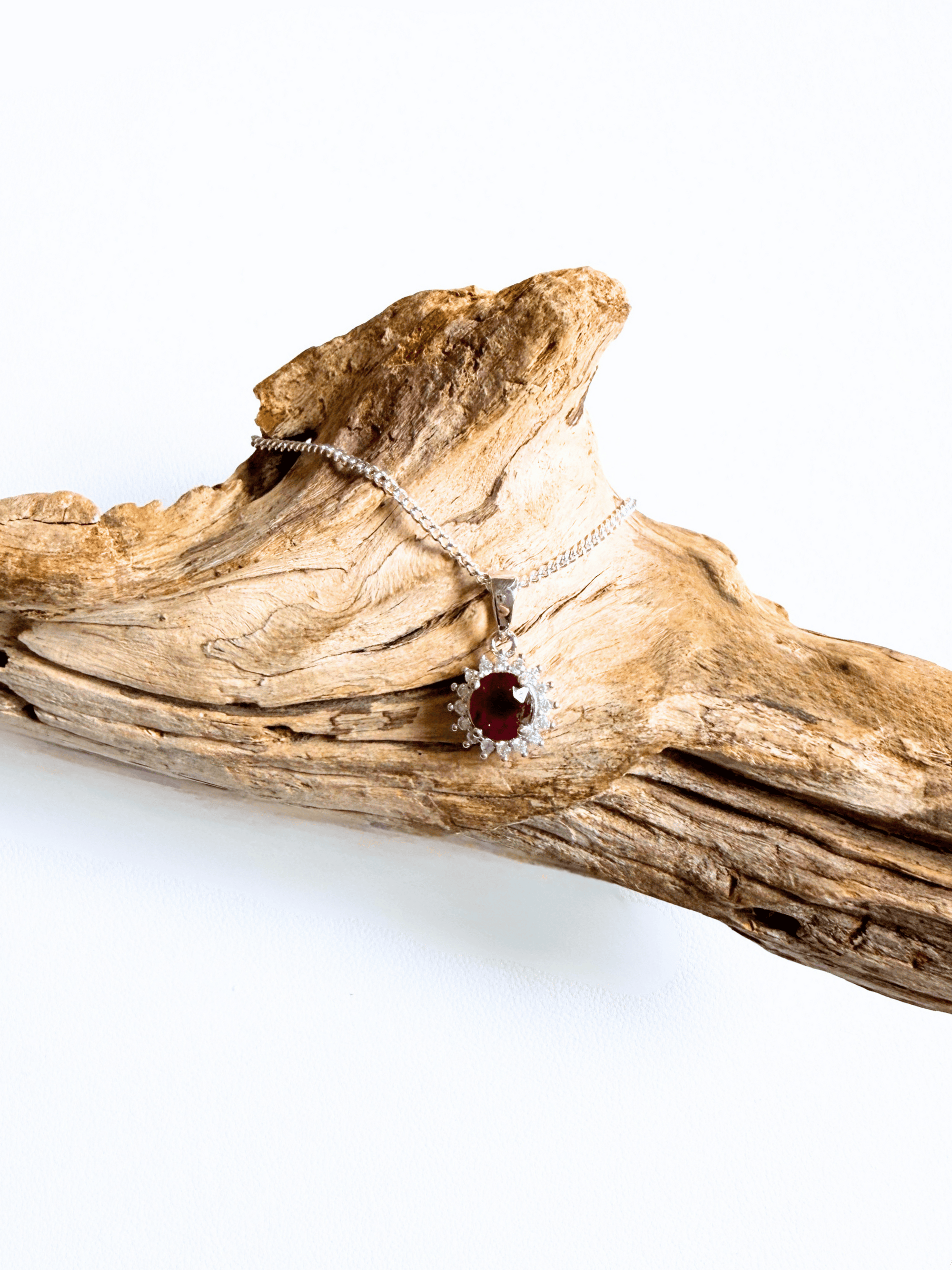 A silver chain necklace with a halo pendant featuring a round red ruby gemstone, displayed on a piece of natural driftwood against a white background.