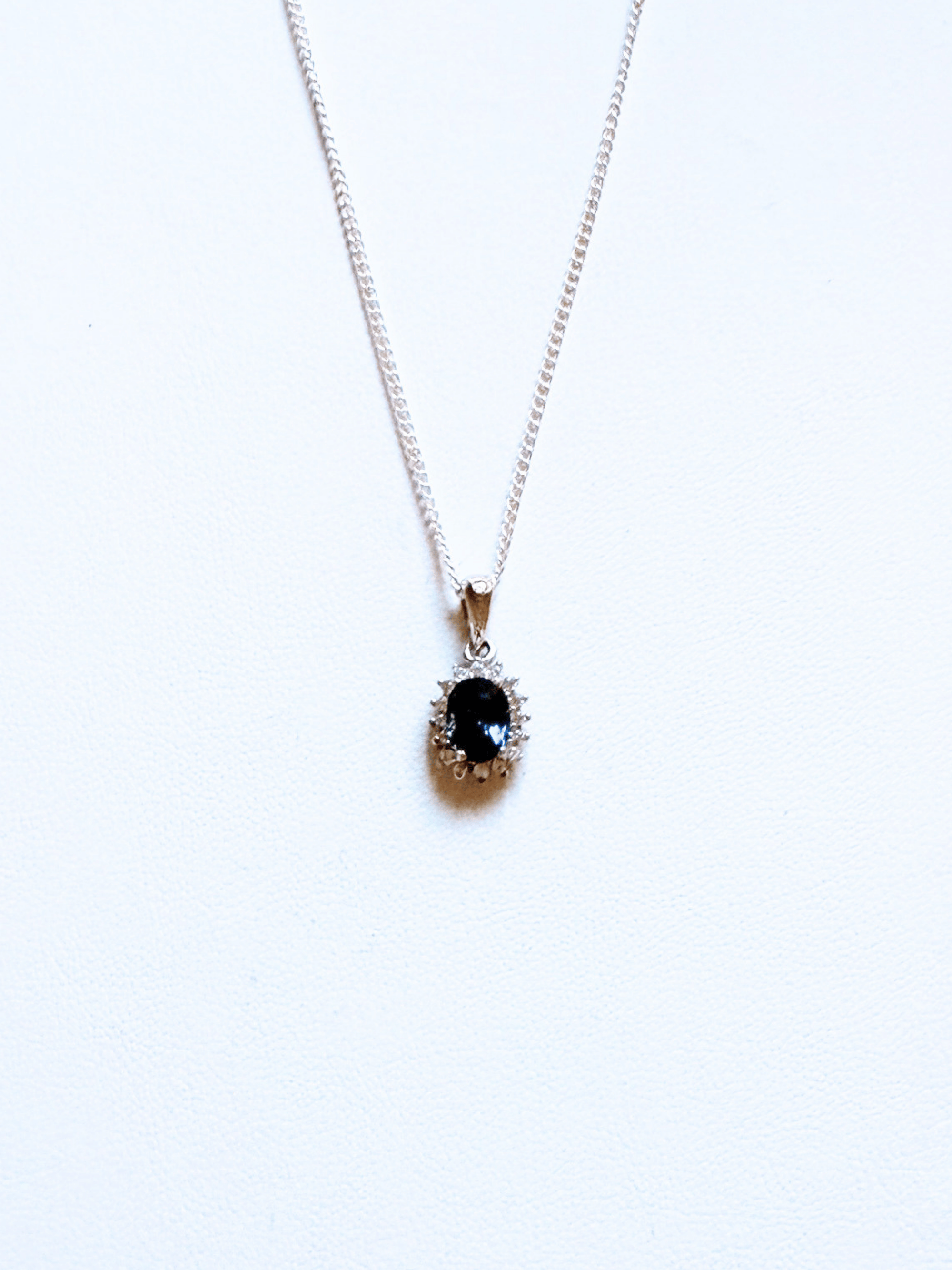 A silver chain necklace with a teardrop-shaped pendant featuring a dark blue oval gemstone surrounded by small clear stones, displayed on a plain white background.