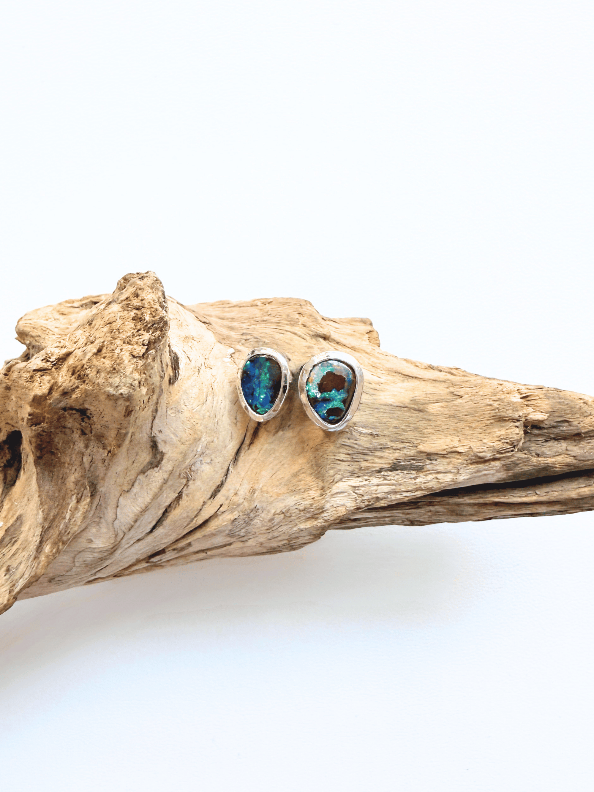 A pair of silver stud earrings with blue-green opal-like stones, displayed on a piece of natural driftwood against a white background.