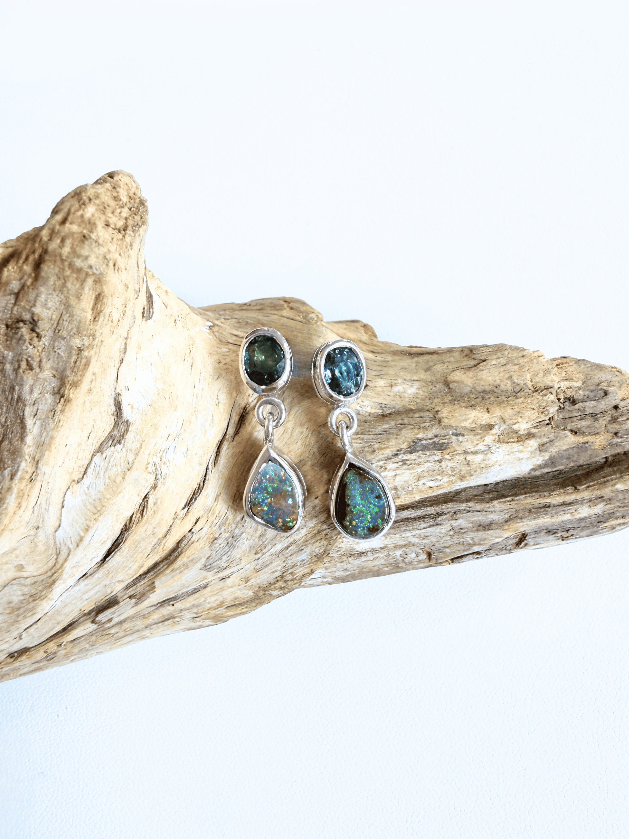 A pair of silver dangle earrings with round and teardrop-shaped iridescent green-blue opal stones, displayed on a piece of natural driftwood against a white background.