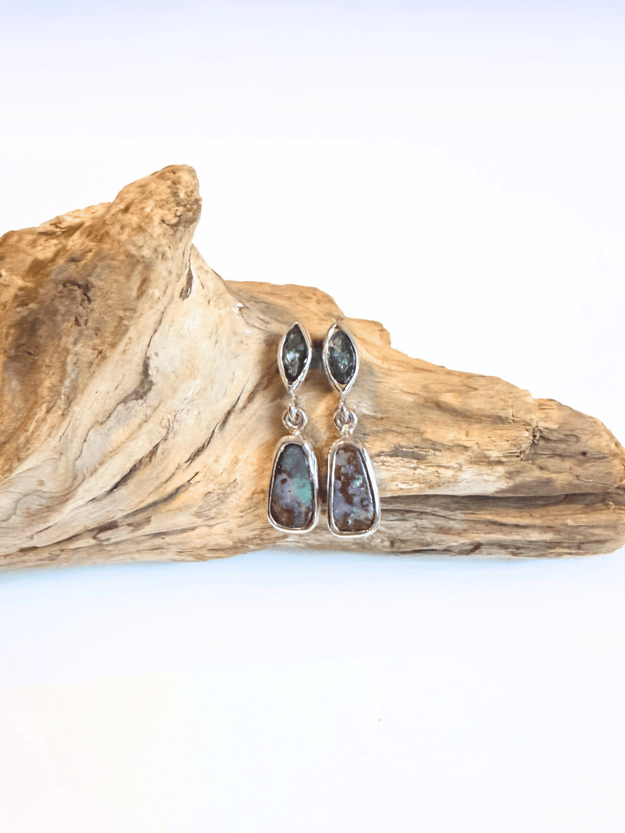 A pair of silver drop earrings with marquise-shaped dark green stones at the top and teardrop-shaped opals at the bottom, displayed on a piece of natural driftwood against a white background.