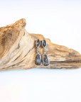 A pair of silver drop earrings with marquise-shaped dark green stones at the top and teardrop-shaped opals at the bottom, displayed on a piece of natural driftwood against a white background.