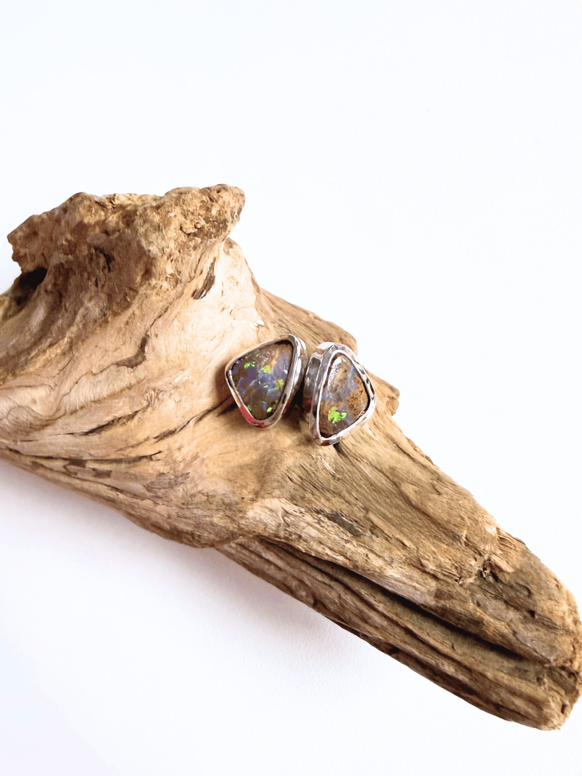 A pair of triangular stud earrings with iridescent opal-like stones in silver settings, displayed on a piece of natural driftwood against a white background.