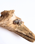 A pair of triangular stud earrings with iridescent opal-like stones in silver settings, displayed on a piece of natural driftwood against a white background.
