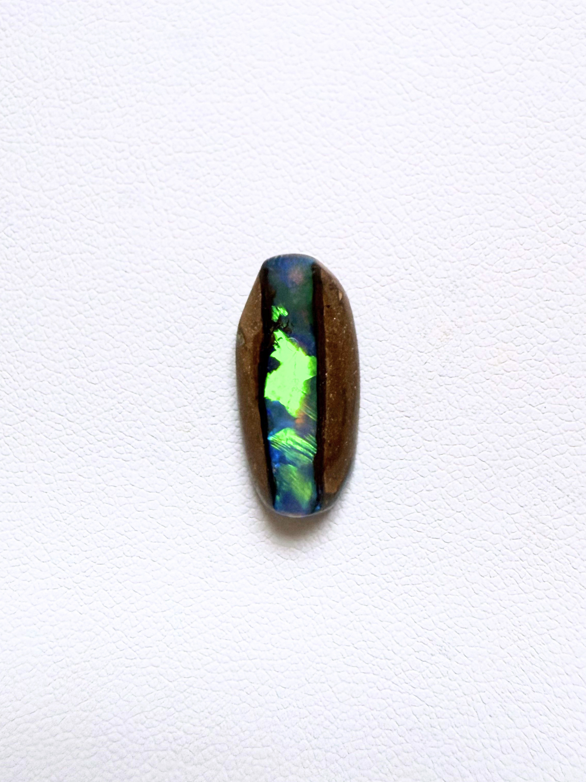 A polished oval cabochon gemstone showing vibrant iridescent play-of-color with green, blue, and purple hues, set in a natural brown opal matrix, displayed on a clean white textured background.