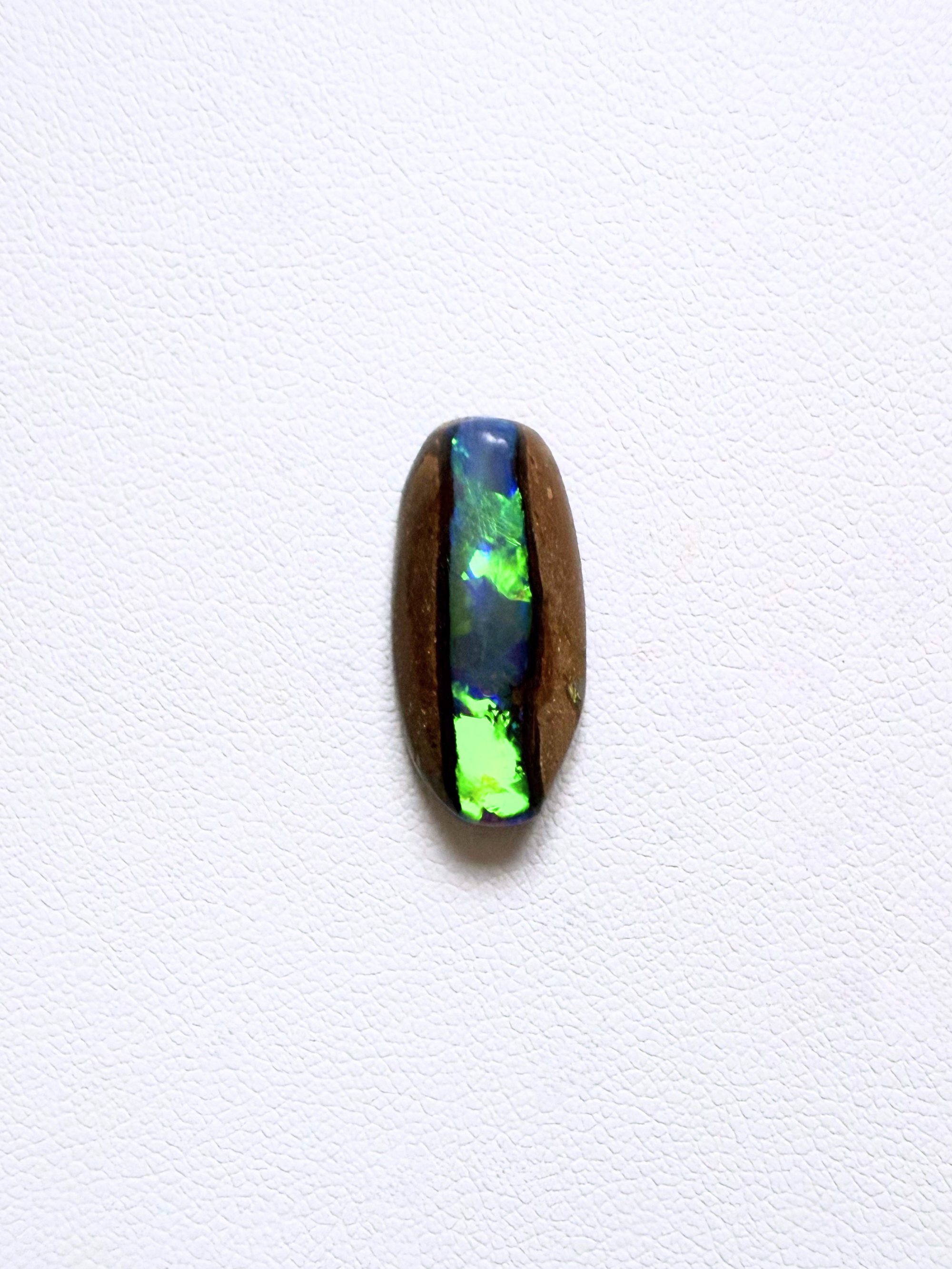 A polished oval cabochon gemstone featuring a vibrant green and blue opal inlay set on a dark brown wooden backing, displayed against a clean white textured background.