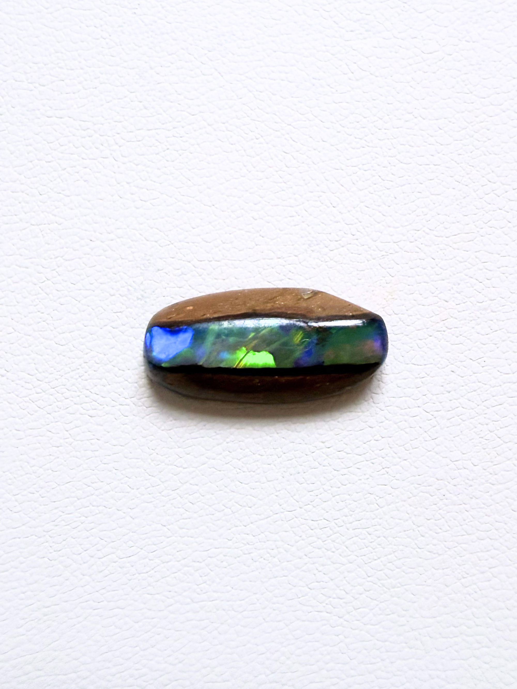 A polished oval cabochon pipe opal featuring a natural brown outer layer and a vibrant iridescent blue, green, and purple opal inlay, displayed on a textured white background.