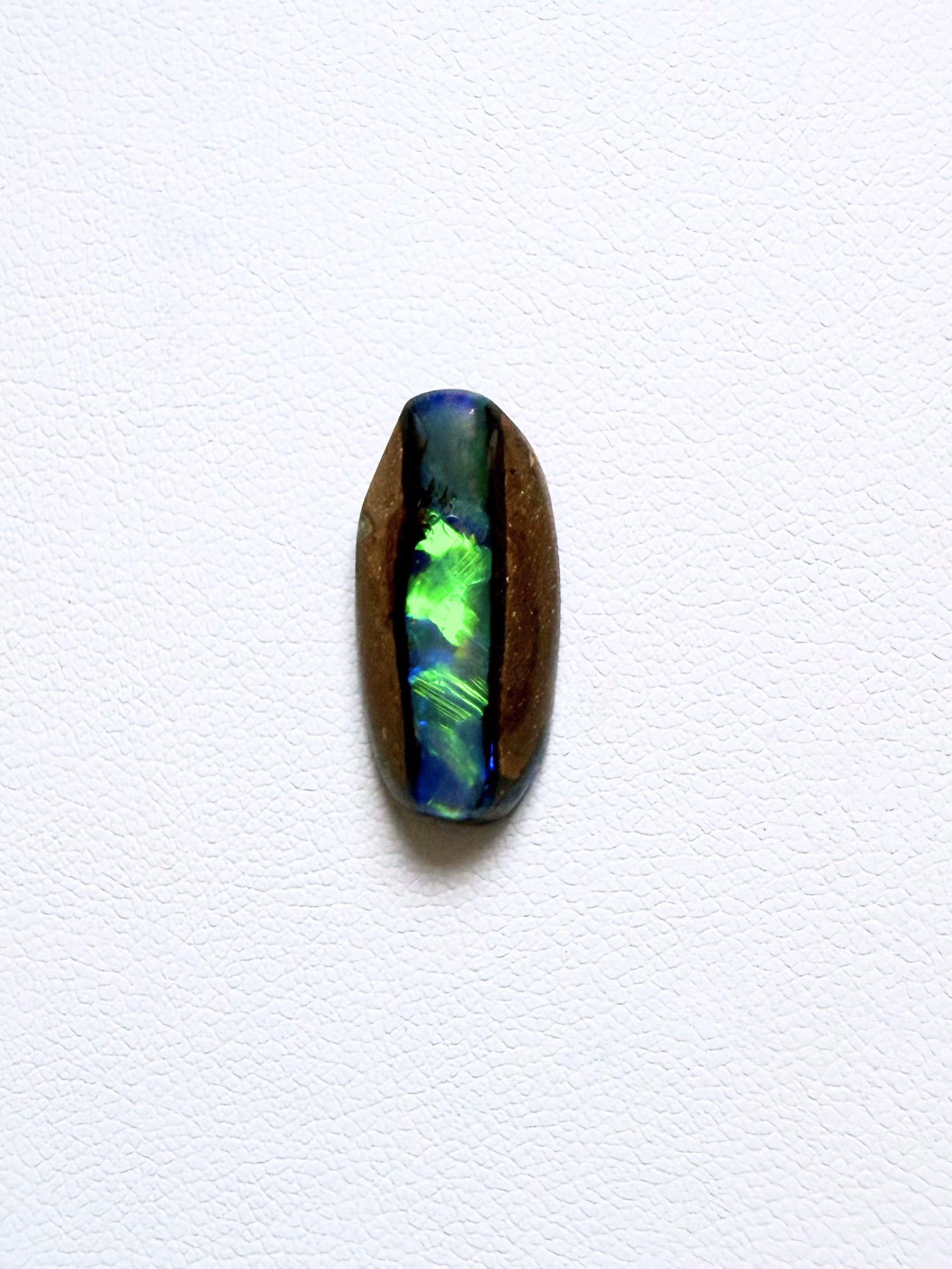 A polished oval cabochon pipe opal showing vibrant iridescent play-of-color with green, blue, and purple hues, set in a dark brown bezel on a textured white background.