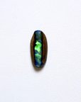 A polished oval cabochon pipe opal showing vibrant iridescent play-of-color with green, blue, and purple hues, set in a dark brown bezel on a textured white background.